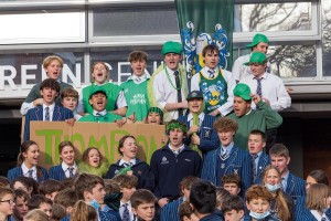 Students in Thompson House are pictured in uniform with green accessories as they stand, chanting and holding a cardboard sign that reads 'Thompson'.